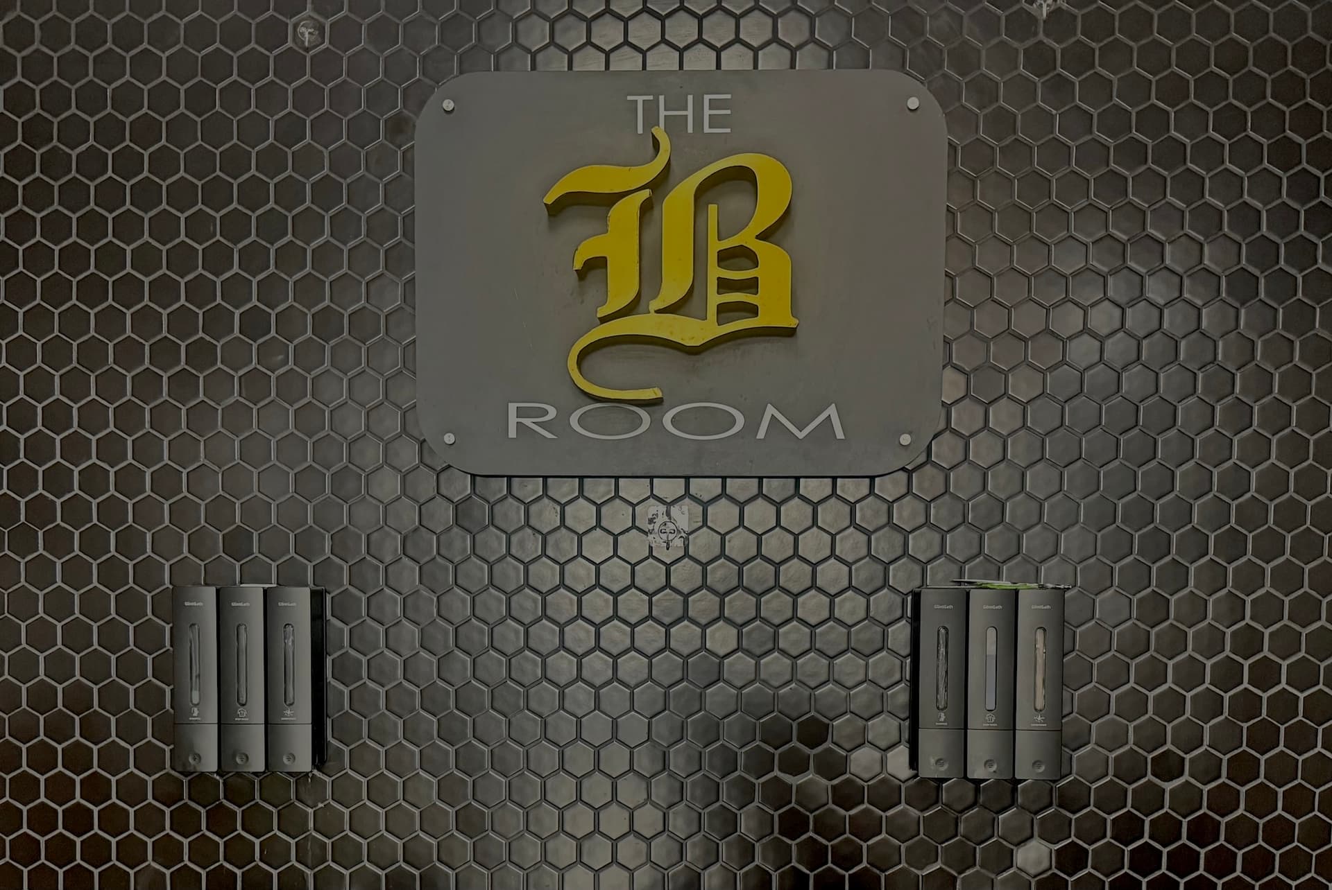 Modern barbershop wall featuring gold B Room logo on gray hexagonal tile with chrome dispensers 1