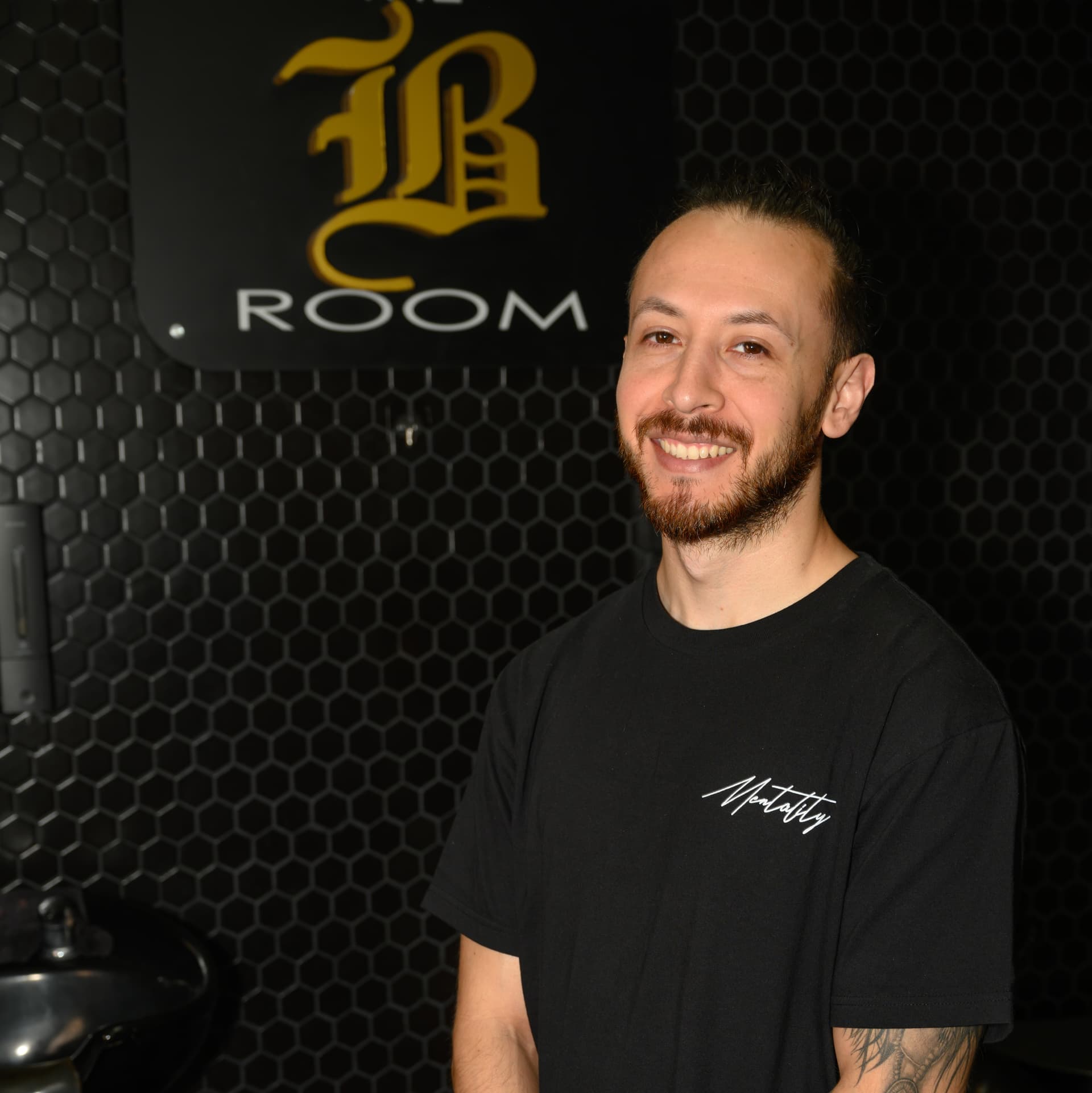 Michael, barber smiling in black shirt, standing in front of B Room sign with hexagonal wall pattern